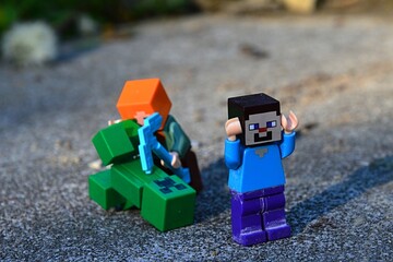 Naklejka premium LEGO Minecraft Steve figure running from Alex with diamond pickaxe fighting with explosive green Creeper mob monster on large stone block. 