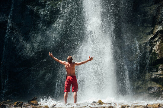 Middle-aged man dressed only red trekking shorts standing under the mountain river waterfall, rose arms up and enjoying the splashing Nature power. Traveling, trekking and nature concept image.