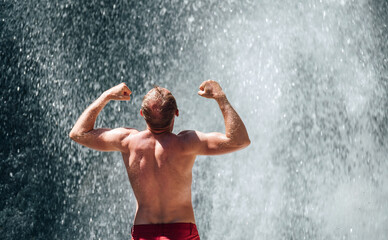 Middle-aged topless man standing under the mountain river waterfall rose arms up showing biceps and enjoying the splashing Nature power. Fit people, trekking and natural beauty concept image.