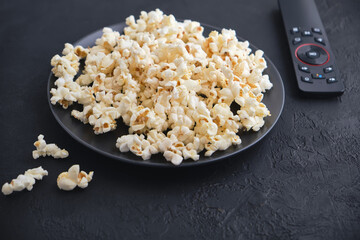 Black plate of popcorn with salted, remote control TV on a black table.