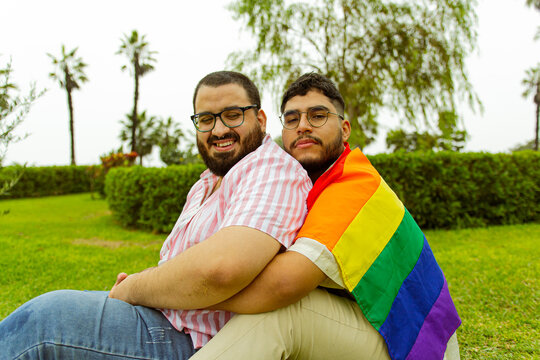 Retrato frontal de pareja de novios gay con barba abraz&aacute;ndose en el parque con una bandera LGBTQ 