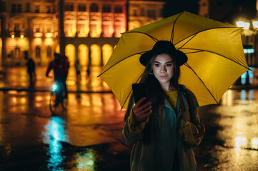 Young beautiful woman using a smartphone and holding a yellow umbrella during night
