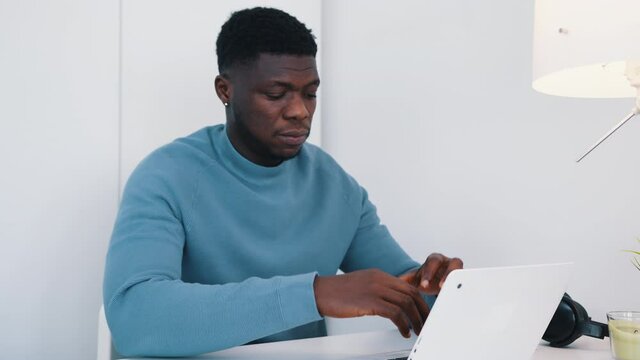 African American black man sitting at the table in front of a laptop working. Stressed out man working on his laptop. Work stress or work load concept. Headphones and a table lamp can be seen. 