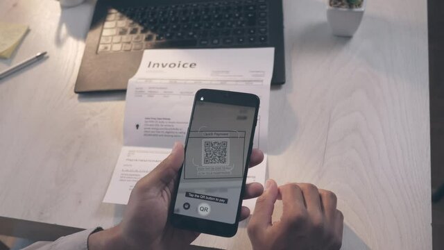 POV shot of an African-American man sitting at the desk, holding a smartphone in the left hand, bringing it closer to an invoice, scanning QR code and paying with a credit card mobile app