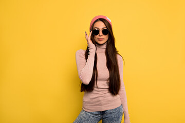 Fototapeta premium Elegant european girl with long dark hair wearing black sunglasses touching her glasses and posing before yellow wall