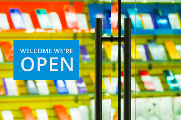 Mobile phone store showcase, view through the glass front door. Welcome We're Open sign hanging on a glass entrance door of a shop selling electronic gadgets