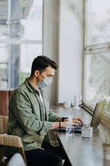 young woman sitting in a mask at coffee shop working on laptop, using disinfectant