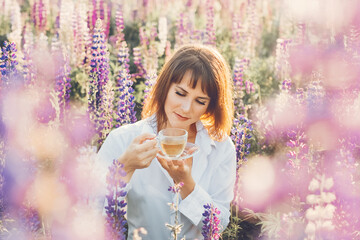 girl in white shirt with in field of lupins and holds glass cup of floral tea in summer