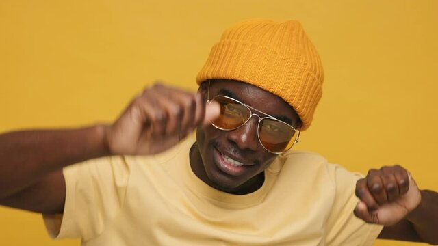 Dancing happy funny black African American man moving to music waving his hands up and down smiling with pleasure in yellow hat and sunglasses on yellow background. Positive emotions of people life