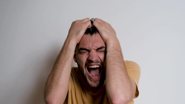 Young handsome European Caucasian man with beard shouts no and grabs his head with both hands. Person depicts the emotions of anger on white background.