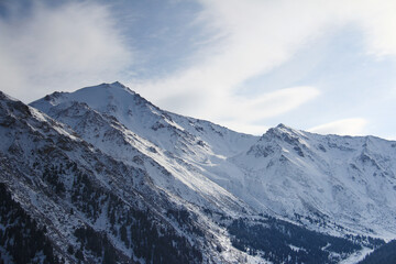 Snow-covered slopes and rocky peaks of the Alatau mountain range, trees grow on the slopes, the sky with clouds, winter, sunny