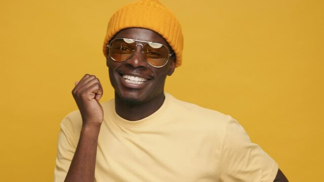 Dancing happy funny black African American man moving to music waving his hands up and down smiling with pleasure in yellow hat and sunglasses on yellow background. Positive emotions of people life
