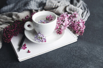 Spring romantic background with lilac flowers in a white cup, with a notebook and a pen for drawing on a background of delicate draped fabric on a gray table, closeup