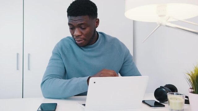 African American black man sitting at the table in front of a laptop. Stressed out man working on his laptop and checking his phone. Work stress concept. Headphones and a table lamp can be seen. 
