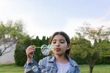 Hispanic girl blowing soap bubbles in the backyard with green grass.