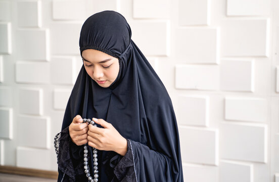 beautiful asian muslim woman looking down smiling happy praying and holding a misbaha bead, wearing black hijab robe, sitting in private praying room with white brick and bright light background