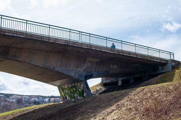 An underneath view of a cement overpass bridge with metal rails. The highway structure has grass on the banks and a walking trail is underneath. There's a blue sky with trees in the background. 