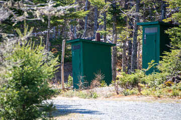 Two green wooden outhouses or bathrooms hidden in the woods at a campground and park. There's a gravel road leading to the privy buildings. The evergreen trees in the park are lush green with sun. 
