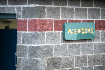 A washroom sign with yellow letters on a green background in a park. The sign is green in color. The wall is grey brick with a red color brick stripe. There's a black door with a silver metal handle. 