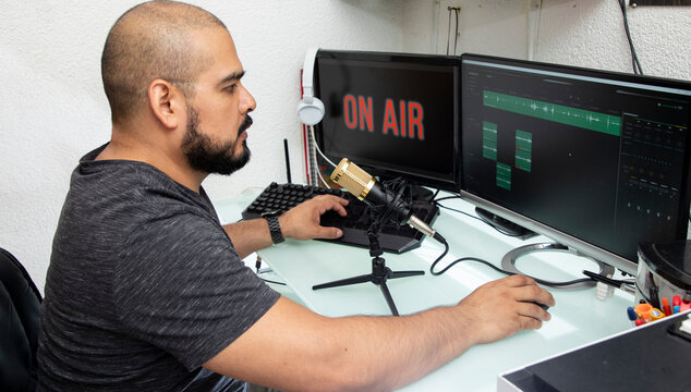 Hombre latino haciendo una transmisi&oacute;n en line&aacute; de un podcast, usando un micr&oacute;fono y su ordenador