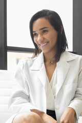 detail of a young latin female doctor with short hair sitting, smiling and wearing her white uniform coat, health professional at her workplace