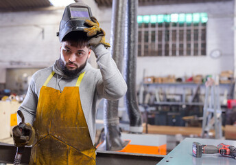 Portrait of young skilled welder wearing protective apron, gloves and helmet standing in...