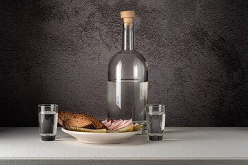 A glass bottle with glasses, with a strong drink and a snack on a white plate, pickles, bacon, bread, on a white countertop against a gray wall