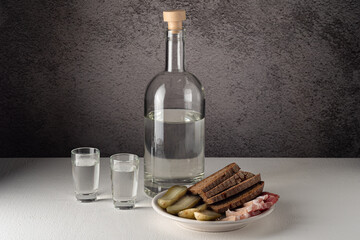 A glass bottle with glasses, with a strong drink and a snack on a white plate, pickles, bacon, bread, on a white countertop against a gray wall