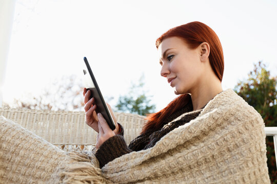 Young woman with red hair reading digital tablet on porch - Powered by Adobe
