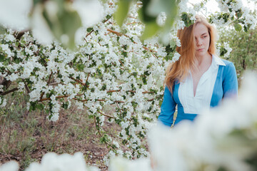 A woman stands sadly among the beautiful blooming gardens. Flowers partially cover the foreground. Apple trees are blooming. The blonde is wearing a strict blue dress with a white collar