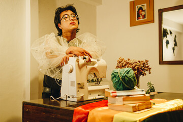 Joven queer diseñador de modas con lentes posando junto a su máquina de coser 