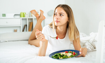 Obraz na płótnie Canvas Cheerful young girl in lingerie having a salad while laying in the bed. High quality photo
