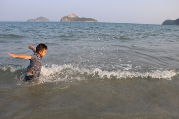 Fototapeta na wymiar Thailand-February26,2021 : Cute Asian boy, good mood, charming, wearing plaid shirt. Standing playing in the water Splashing water on the sea with fun and joy Behind the sky and an island in the sea.