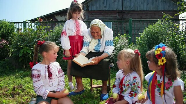 An old grandmother is sitting on the street and reading a book. She is dressed in national ukrainian clothing vyshyvanka. Girls listen carefully to their grandmothers who read them fairy tales
