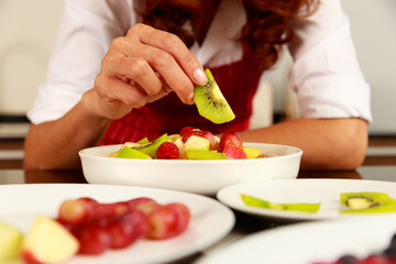 Obraz na płótnie Canvas Closeup shot of chef hand putting kiwi slice into tasty yummy delicious sweet mixed fruit salad white bowl on kitchen table behind raw fresh cut apple and grape material dishes in blurred foreground