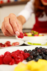 Obraz na płótnie Canvas Closeup shot of chef hand putting raspberry into yummy sweet mixed fruit salad in white bowl on kitchen table. Organic and natural food concept.