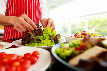 Closeup shot of houswife hand decorating green mixed vegetable salad on countertop desk in kitchen with raw fresh organic material in blurred foreground