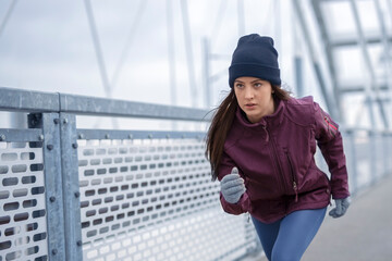 Focused young fitness woman in winter sportswear, winter skully hat and winter gloves jog on bridge during cold day
