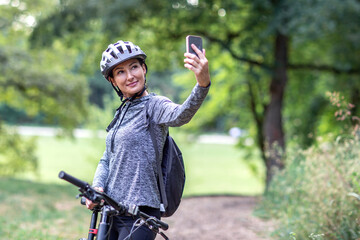 Naklejka premium Young smiling woman in a tracksuit on a mountain bike take selfie with smartphone in the park