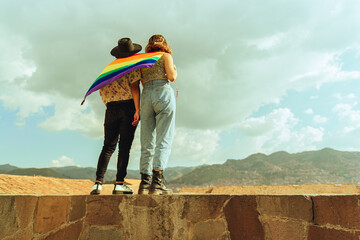 Amigos queer y bisexual portando la bandera LGTBQ en sus espaldas mientras observan la ciudad de...