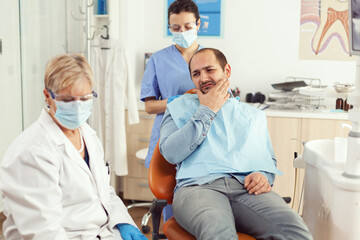 Fototapeta premium Man patient putting his hand on cheek showing toothache complaining about tooth pain. Senior woman explaining dental problem to doctor indicating mouth while sitting on stomatological chair