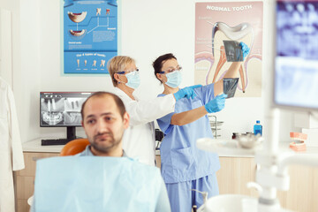 Obraz premium Sick man patient waiting for teeth examination sitting on dental chair in stomatology clinic room. Stomatologist doctor and medical nurse preparing for orthodontic surgery while examining radiography