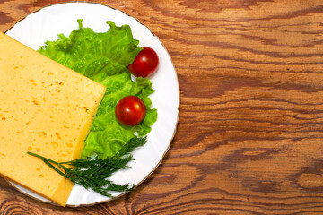 a large piece of cheese lettuce cherry tomatoes and herbs on a white plate on a wooden table copy space