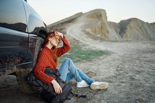 a traveler in a sweater and jeans sits on the ground with a backpack in her hand near a car in the mountains