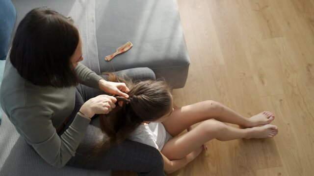 Mom braids her daughter's pigtail. A woman on the couch weaves a braid for a teenage girl, a schoolgirl.
