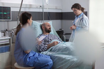 Medical team monitoring sick man explaining disease symptom examining medical recovery in hospital ward. Patient sitting in bed waiting specialist treatment during rehabilitation appointment