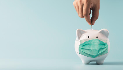 Hand putting coin to white piggy bank saving which wearing surgical mask on blue background and...