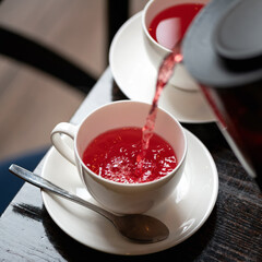 Red tea from frozen berries. Hot drink pouring from teapot into white cup and saucer on wooden table. Tea time. Close up shot. Soft focus.