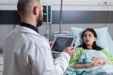 Practitioner man doctor explaining disease expertise to sick woman using tablet computer. Patient sitting in bed recovering after medical surgery in hospital ward while black nurse arrange bed