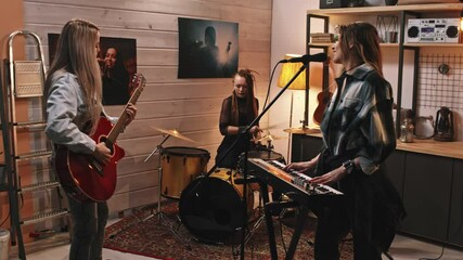 Medium slowmo of young energetic all-girl music band consisting of drummer, guitarist and pianist playing music and singing in retro-style studio with posters of famous musicians on walls - Powered by Adobe